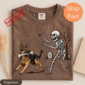 Puede incluir: Una camiseta marrón de cuello redondo con un gráfico de un perro pastor alemán corriendo con un hueso en la boca mientras es perseguido por un esqueleto en un cementerio. La camiseta tiene una etiqueta de la marca Comfort Colors en el cuello.