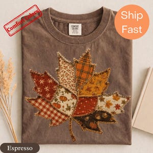 May include: An espresso-colored t-shirt featuring a large, central maple leaf graphic composed of various fabric-style patches. The patches display patterns including orange plaid, white pumpkins on a cream background, red gingham, and brown acorns.