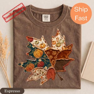 Peut inclure: Un t-shirt marron à manches courtes avec un grand motif central en forme de feuille d'érable composé de divers empiècements. Les motifs présentent des citrouilles, des vignes et des feuilles d'automne dans des tons orange, bleu sarcelle et marron.