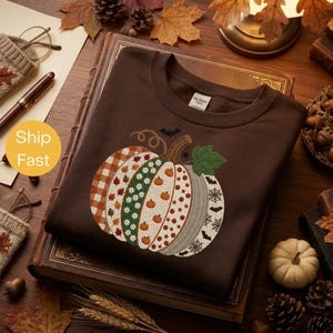 Peut inclure: Un sweat-shirt à col rond marron foncé orné d'un grand motif central représentant une citrouille divisée en segments verticaux avec divers motifs, dont du tartan orange, des feuilles d'automne, des marguerites et des toiles d'araignée.