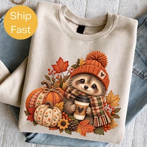 May include: A beige crewneck sweatshirt featuring a graphic of a hedgehog wearing an orange knit beanie and a plaid scarf. The hedgehog holds a coffee cup with a heart design, surrounded by pumpkins, autumn leaves, berries, and a pinecone.