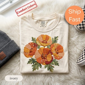 Peut inclure: Un t-shirt ivoire arborant un motif central composé de quatre fleurs stylisées dans des tons orange, jaune et marron. Le design floral est constitué de divers motifs de tissus, incluant du vichy et des imprimés floraux, avec des feuilles vertes.