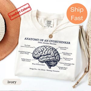 May include: An ivory t-shirt featuring a black anatomical brain illustration with the text Anatomy of an Overthinker, Homo Anxietus Maximus, and labels pointing to various parts of the brain including The Vault and Constant Worry Generator.
