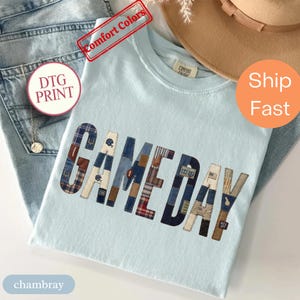 Könnte beinhalten: Ein hellblaues T-Shirt mit dem Wort GAME DAY in großen Buchstaben. Jeder Buchstabe ist mit einem Patchwork aus verschiedenen Stoffmustern gefüllt, darunter Schottenkaro, Denim und braune Texturen, mit integrierten Football-Symbolen.
