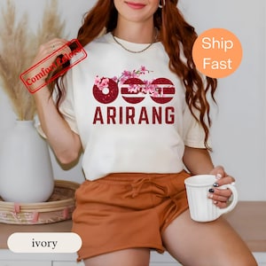 Könnte beinhalten: Ein elfenbeinfarbenes Kurzarm-T-Shirt mit einem Grafikdesign aus drei dunkelroten Glitzerkreisen und rosa Kirschblütenzweigen. Der Text ARIRANG ist in dunkelroten Glitzer-Großbuchstaben unter den Kreisen aufgedruckt.
