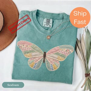 Puede incluir: Una camiseta verde espuma de mar con un gran gráfico de mariposa compuesto por varios parches de tela en tonos pastel con costuras visibles. La camiseta tiene un cuello redondo y una etiqueta de la marca Comfort Colors en el interior.