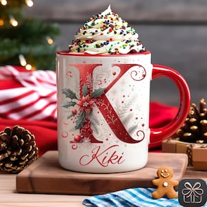 May include: A white ceramic mug with a red handle, featuring a large red letter K decorated with poinsettias, holly leaves, and berries. The name Kiki is printed in red script below the letter. The mug is filled with whipped cream and sprinkles.