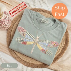Puede incluir: Una camiseta de cuello redondo en color verde salvia con un gráfico grande de una libélula en la parte delantera. Las alas de la libélula están compuestas por varios patrones de tela de patchwork, incluyendo flores, cuadros vichy y tonos sólidos en rosa, azul, amarillo y verde.