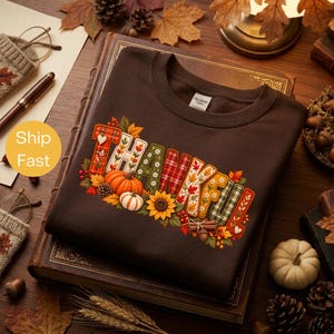 Pode incluir: Uma camisola de gola redonda castanho-escura com a palavra THANKFUL em letras grandes com vários padrões. O design inclui elementos temáticos de outono como abóboras, girassóis, pinhas, bagas e paus de canela.