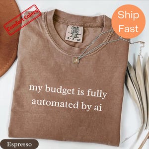 Może przedstawiać: Koszulka z krótkim rękawem w kolorze espresso z okrągłym dekoltem. Na środku klatki piersiowej znajduje się biały nadruk z tekstem 'my budget is fully automated by ai'. Na tkaninie spoczywa srebrzysty naszyjnik z małą okrągłą zawieszką.