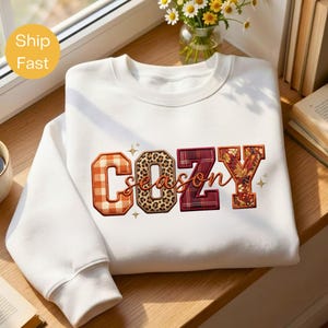 Puede incluir: Una sudadera blanca de manga larga con la palabra COZY en letras grandes con patrones variados que incluyen cuadros naranjas, estampado de leopardo, cuadros rojos y hojas de otoño, junto a la palabra season en letra cursiva.