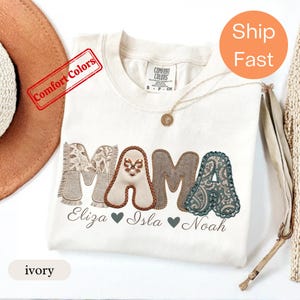 Puede incluir: Una camiseta color marfil con la palabra MAMA en letras grandes de tela texturizada con varios patrones. La primera M tiene un estampado floral, la A un pequeño lazo, la segunda M un patrón de espiga y la última A un estampado de cachemira. Debajo, los nombres Eliza, Isla y Noah están impresos en cursiva con corazones.