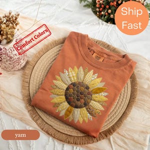 May include: A rust-coloured crewneck t-shirt featuring a large sunflower graphic on the front. The sunflower petals are composed of various yellow, gold, and tan fabric patterns, while the centre consists of small, dark brown circular patches.