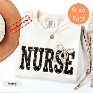 Puede incluir: Una camiseta de color marfil con la palabra NURSE en letras negras de bloque con puntos blancos. La letra E está decorada con un lazo bordado de color claro. La camiseta tiene cuello redondo y una etiqueta de la marca Comfort Colors.