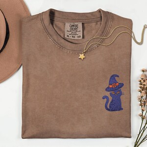May include: A brown crewneck t-shirt featuring an embroidered purple cat wearing a pointed orange-accented witch hat on the lower right side. The shirt has a Comfort Colors label at the inner neckline and is paired with a gold star pendant necklace.