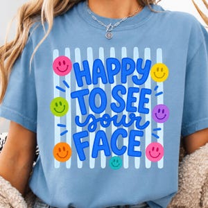 May include: A light blue t-shirt with the phrase "HAPPY TO SEE your FACE" in blue, surrounded by colorful smiley faces. The shirt has a crew neck and short sleeves. The design includes vertical white stripes.