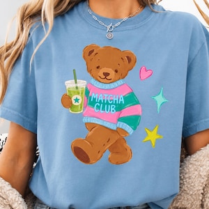 Puede incluir: Camiseta azul claro con un oso de peluche marrón que lleva un suéter rosa y verde con el texto "Matcha Club". El oso sostiene una bebida verde con una estrella, rodeado de un corazón rosa y estrellas.