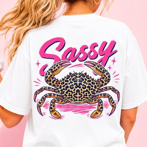 Op de afbeelding: Een wit t-shirt met een afbeelding op de achterkant van een krab met een bruin en zwart luipaardpatroon. Boven de krab staat het woord Sassy gedrukt in een vet, felroze scriptlettertype. Het ontwerp bevat kleine roze steraccenten.