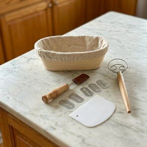 May include: A bread-making kit with a banneton proofing basket, dough whisk, lame with blades, and a dough scraper. The basket is beige with a linen liner. The tools have wooden handles and are arranged on a marble countertop.