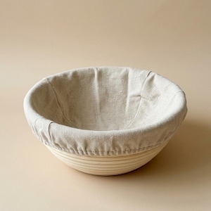 May include: A round banneton proofing basket in a light colour, with a linen liner. The basket has a ribbed exterior and is designed for proofing bread dough. The linen liner is draped inside the basket.