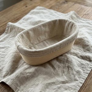 May include: Oval bread proofing basket with a linen liner. The basket is made of light-coloured, woven material. It sits on a beige linen cloth, creating a neutral and natural aesthetic. Ideal for artisan bread baking.