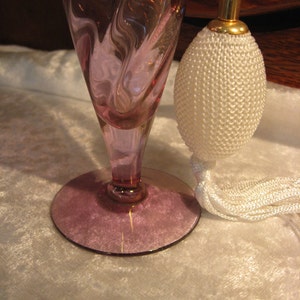 May include: A vintage glass perfume bottle with a swirl design in shades of pink and purple. The bottle has a white tassel and a gold-colored top.