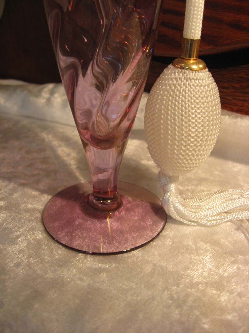 May include: A vintage glass perfume bottle with a swirl design in shades of pink and purple. The bottle has a white tassel and a gold-colored top.