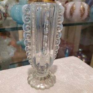 Può includere: Una bottiglia di vetro trasparente con un tappo dorato e un design testurizzato. La bottiglia ha una base a forma di conchiglia e un design decorativo attorno al collo.