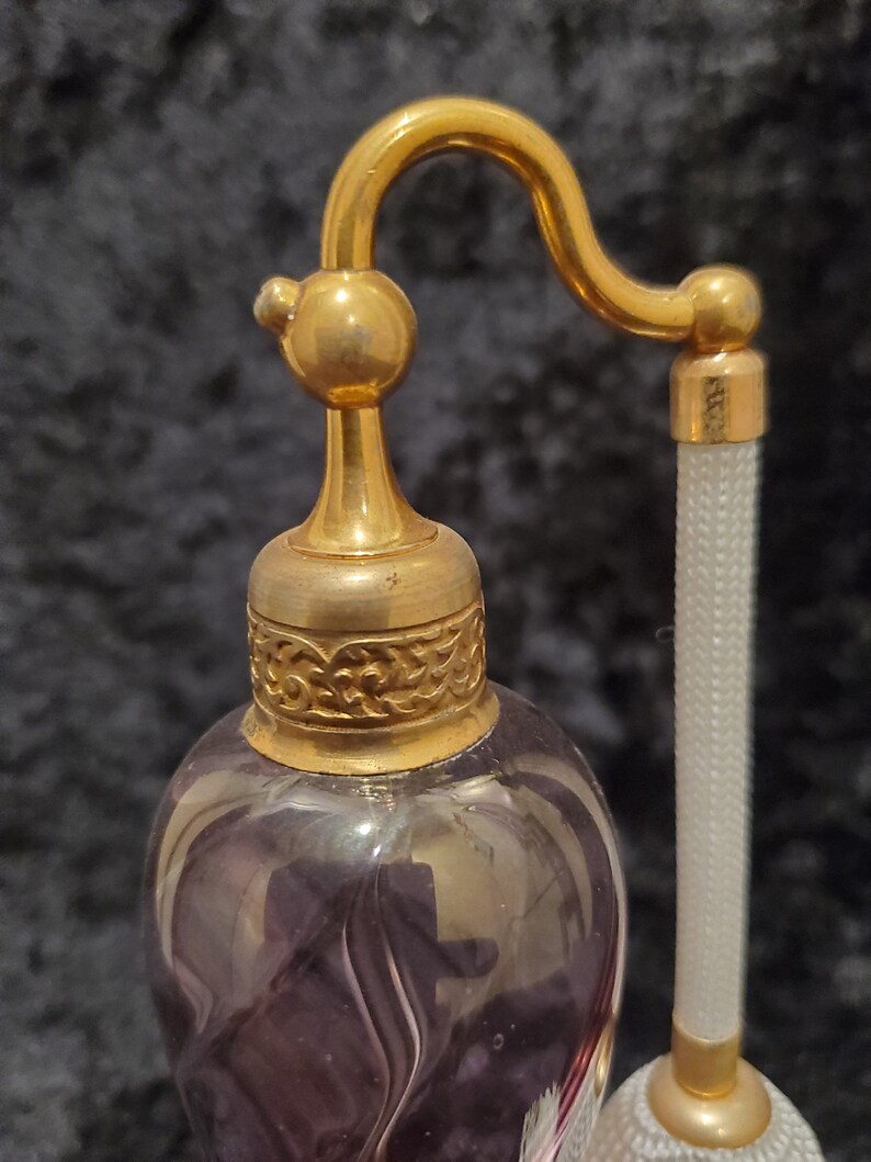 May include: A vintage glass perfume bottle with a gold-toned metal top and a long, thin, white spray nozzle. The bottle is a deep purple color and has a swirling pattern.