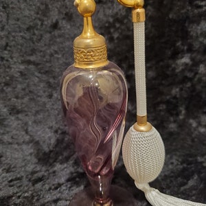 May include: A vintage purple glass perfume bottle with a gold top and a white tassel atomizer. The bottle has a swirl design and sits on a gold base.