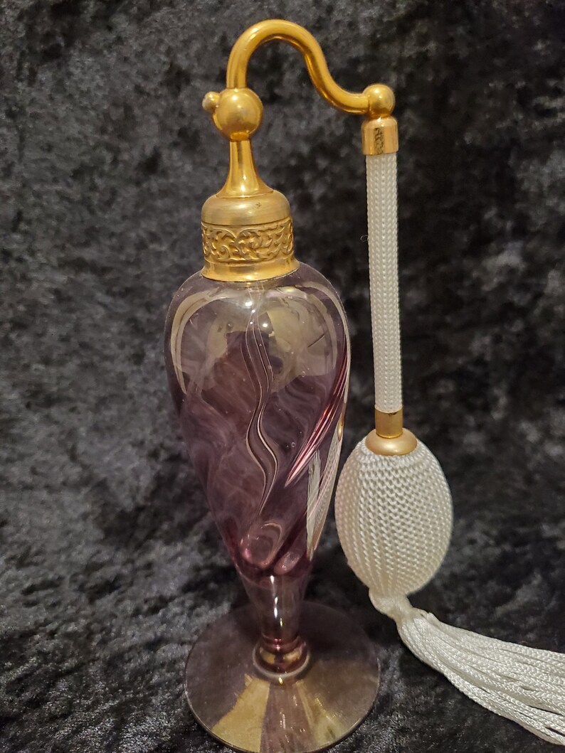 May include: A vintage purple glass perfume bottle with a gold top and a white tassel atomizer. The bottle has a swirl design and sits on a gold base.