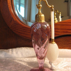 May include: A vintage pink glass perfume bottle with a gold-toned atomizer and a white tassel. The bottle has a swirl design and sits on a white surface.