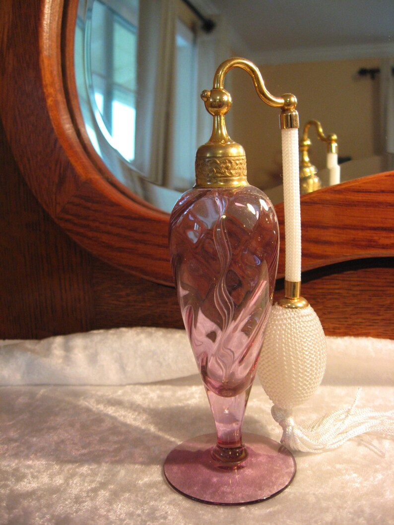 May include: A vintage pink glass perfume bottle with a gold-toned atomizer and a white tassel. The bottle has a swirl design and sits on a white surface.