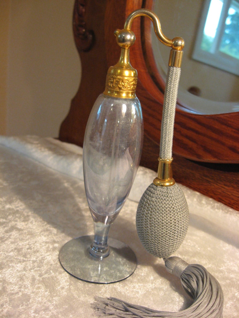 May include: A vintage glass perfume bottle with a gold top and a gray tassel. The bottle has a clear glass body and a silver base.
