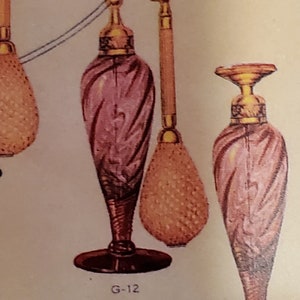 May include: Two pink glass perfume bottles with gold accents and a gold spray nozzle. The bottle on the left has a gold bulb atomizer. The bottle on the right has a black base. The text "G-12" is printed below the bottle on the left.