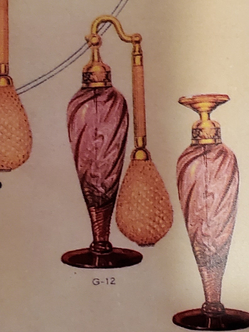 May include: Two pink glass perfume bottles with gold accents and a gold spray nozzle. The bottle on the left has a gold bulb atomizer. The bottle on the right has a black base. The text "G-12" is printed below the bottle on the left.