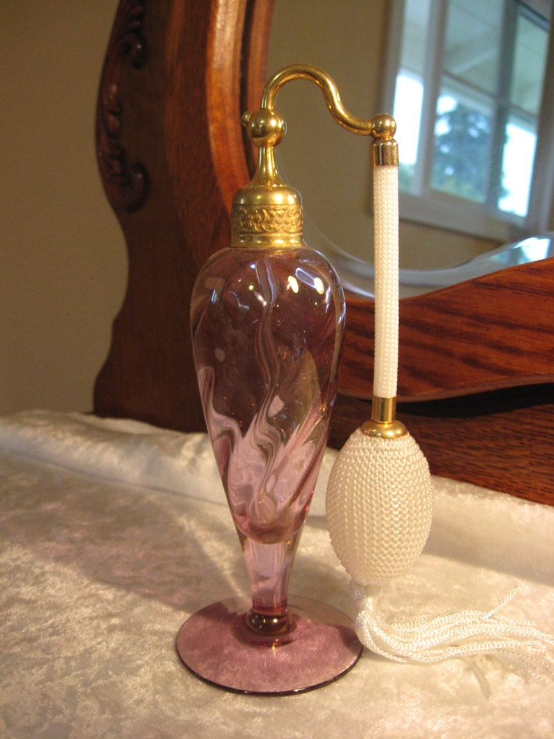 May include: A vintage glass perfume bottle with a swirl design in shades of purple and gold. The bottle has a gold atomizer and a white tassel.