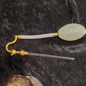 May include: A vintage glass perfume bottle with a gold-colored cap and a white bulb atomizer. The bottle has a purple iridescent color.