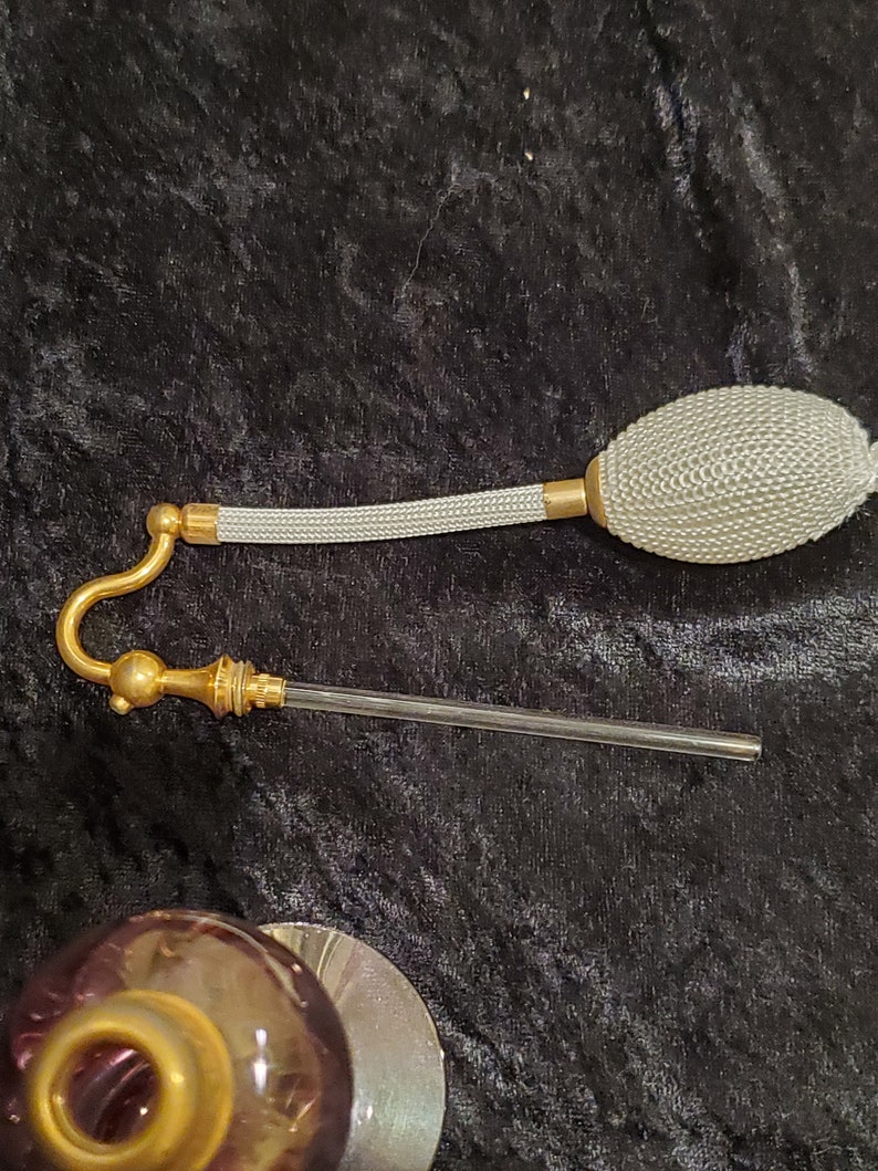May include: A vintage glass perfume bottle with a gold-colored cap and a white bulb atomizer. The bottle has a purple iridescent color.
