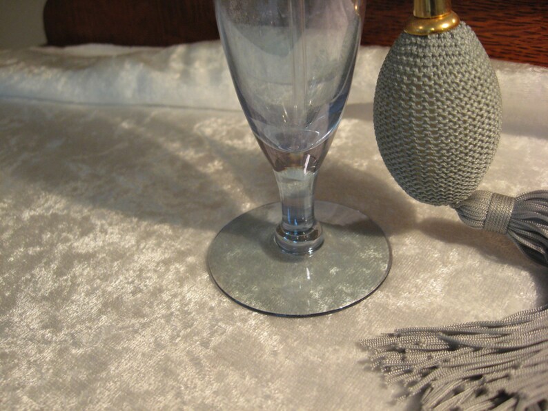 May include: A blue glass perfume bottle with a gray fabric atomizer and tassel. The bottle has a wide base and a narrow neck. The atomizer has a gold top.
