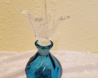 Könnte beinhalten: Eine blaue Glasflasche mit einem durchsichtigen Glasstopfen in Form einer Blume. Die Flasche hat einen runden Körper und einen sechseckigen Boden.