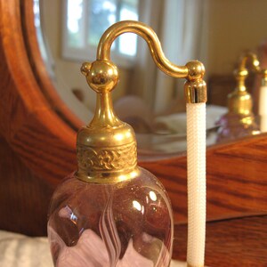 May include: A vintage glass perfume bottle with a gold-toned top and a delicate, swirling design. The bottle has a long, thin neck and a small, round stopper. The bottle is filled with a light purple liquid.