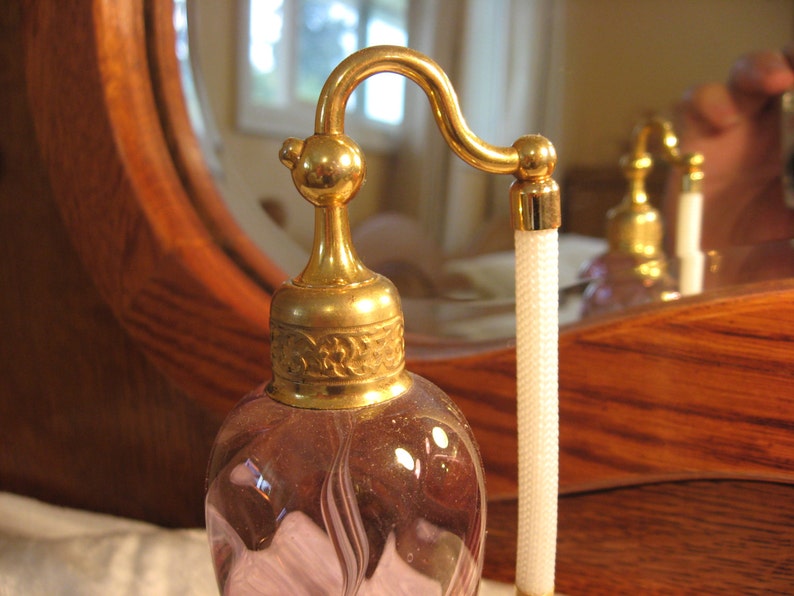 May include: A vintage glass perfume bottle with a gold-toned top and a delicate, swirling design. The bottle has a long, thin neck and a small, round stopper. The bottle is filled with a light purple liquid.