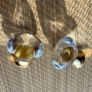 May include: Two circular cabinet knobs featuring clear, faceted glass tops that catch the light, paired with polished gold-toned metal bases. The knobs have a smooth, rounded profile and a reflective surface, showcasing a combination of transparent glass and metallic gold finishes.