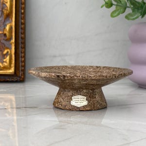 May include: A small, circular, brown speckled cork dish with a wide, shallow bowl and a tapered pedestal base. A white rectangular label on the front of the base features the text "WATER STONE, Genuine Marble, Hand Crafted, 2023".