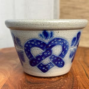 May include: A small, round, light gray ceramic bowl featuring a prominent cobalt blue heart-shaped design on the front. The heart pattern is composed of interconnected loops with small dots, and the bowl has a thick, smooth rim and a tapered base.