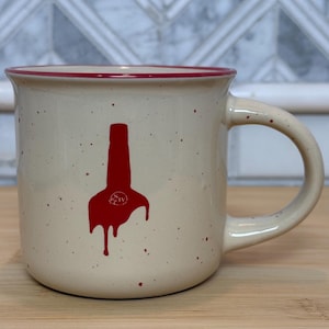 May include: A speckled beige ceramic mug with a red rim and handle. A red graphic of a bottle with dripping paint is on the front. The mug is sitting on a wooden surface.