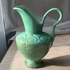 May include: A ceramic pitcher with a glossy mint green glaze and a bulbous body. The vessel features a curved handle, a wide pouring spout, and a pedestal base. Embossed swirl patterns decorate the upper section of the rounded body.