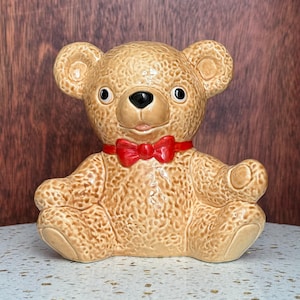 May include: A ceramic teddy bear figurine with a textured tan surface, featuring a glossy finish, round ears, black eyes, a black nose, and a bright red bow tie around its neck. The bear is in a seated position with one arm raised slightly.