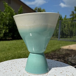 May include: A ceramic goblet with a wide, flared rim and a tapered base. The upper portion of the vessel features a smooth, off-white glaze that transitions into a vibrant, glossy turquoise finish on the lower section and the pedestal base.